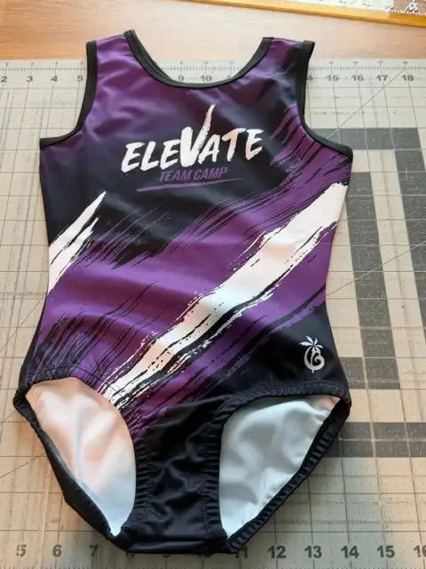 Purple and black athletic leotard with white and pink brushstroke accents, featuring ’elevate team camp’ text across the chest.